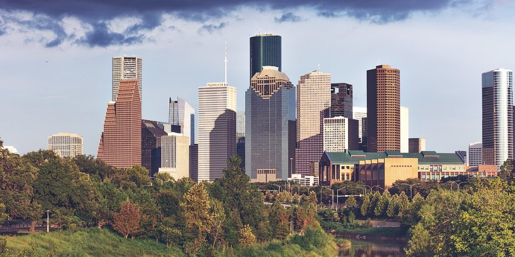 Panoramic view of downtown Houston skyline with tall skyscrapers and green trees in the foreground under a partly cloudy sky.