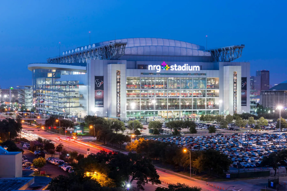 NRG Stadium in Houston, Texas, brightly illuminated at night with a full parking lot and city lights in the background.
