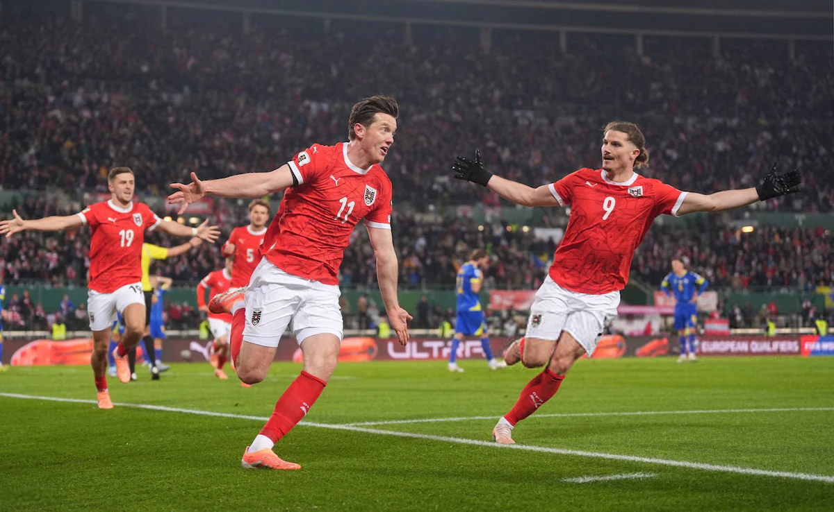 Austria national football team players in red kits celebrate a goal with arms outstretched during a European Championship qualifier, with a cheering crowd in the background.