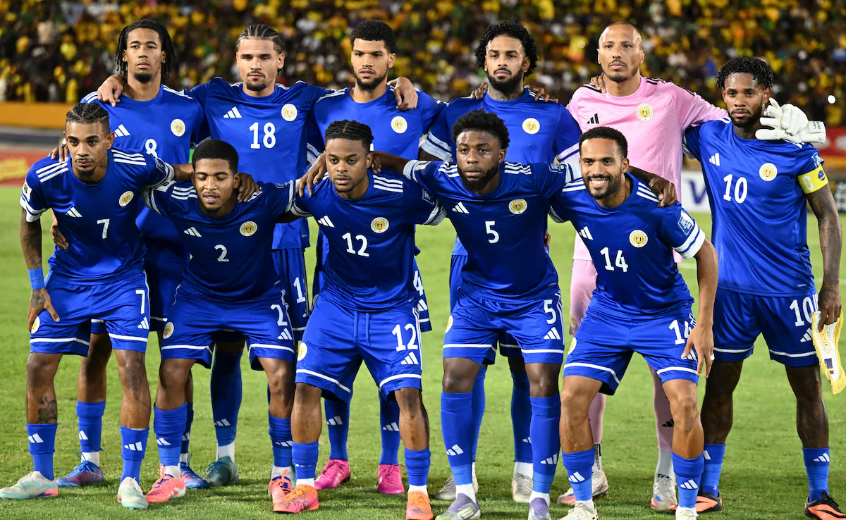 Curaçao national football team in blue uniforms with yellow accents posing for a group photo before a football match, with stadium spectators behind them.