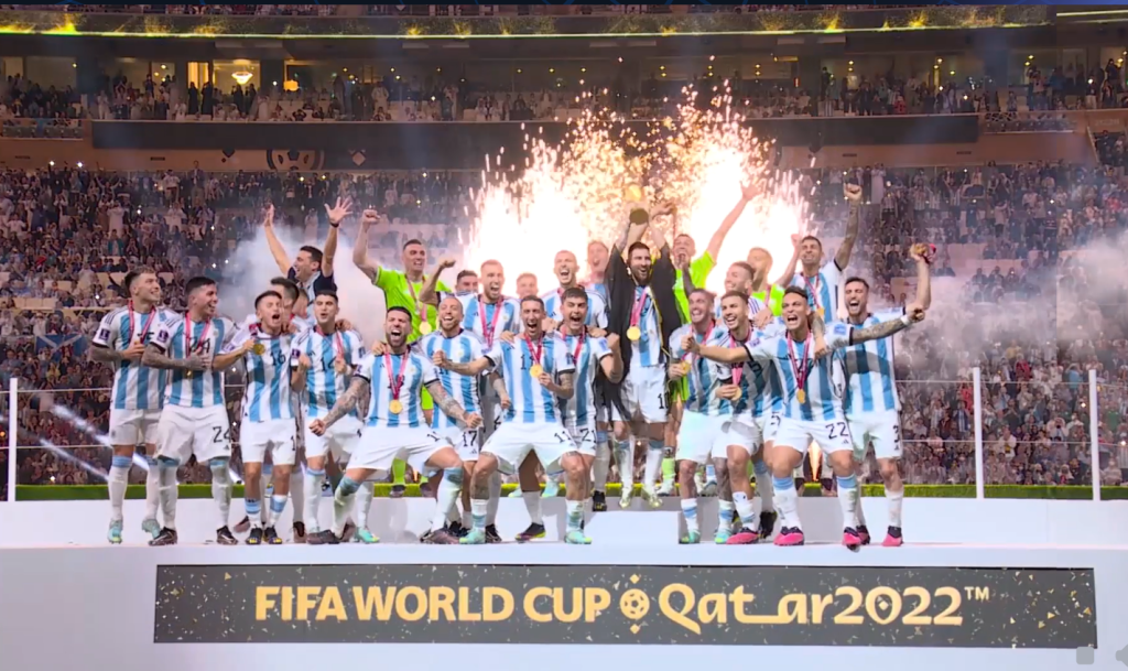 Argentina national football team players celebrating on the podium with the FIFA World Cup trophy, surrounded by fireworks and confetti, after winning the Qatar 2022 tournament.