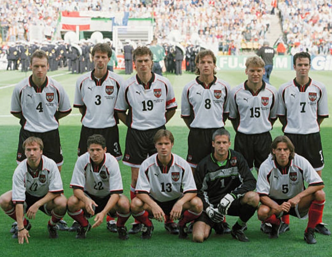 Austria national football team players in white and black kits pose for a group photo on the pitch before a major international football match, with the Austrian flag in the background.