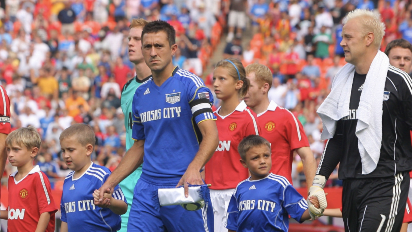 Kansas City Wizards and Manchester United players, along with young mascots, walk onto the field before a soccer match, with a packed stadium in the background.