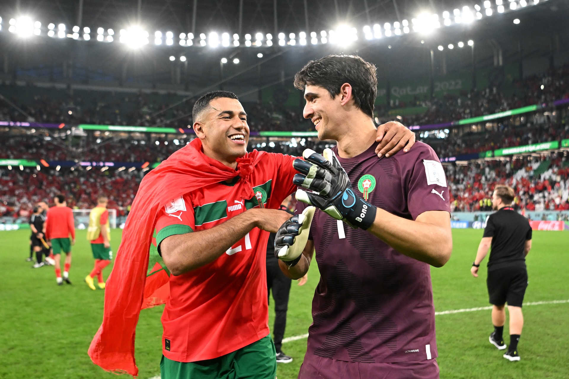 Morocco football players celebrating on the field after a FIFA World Cup match, with one player wearing the national flag and the goalkeeper smiling.