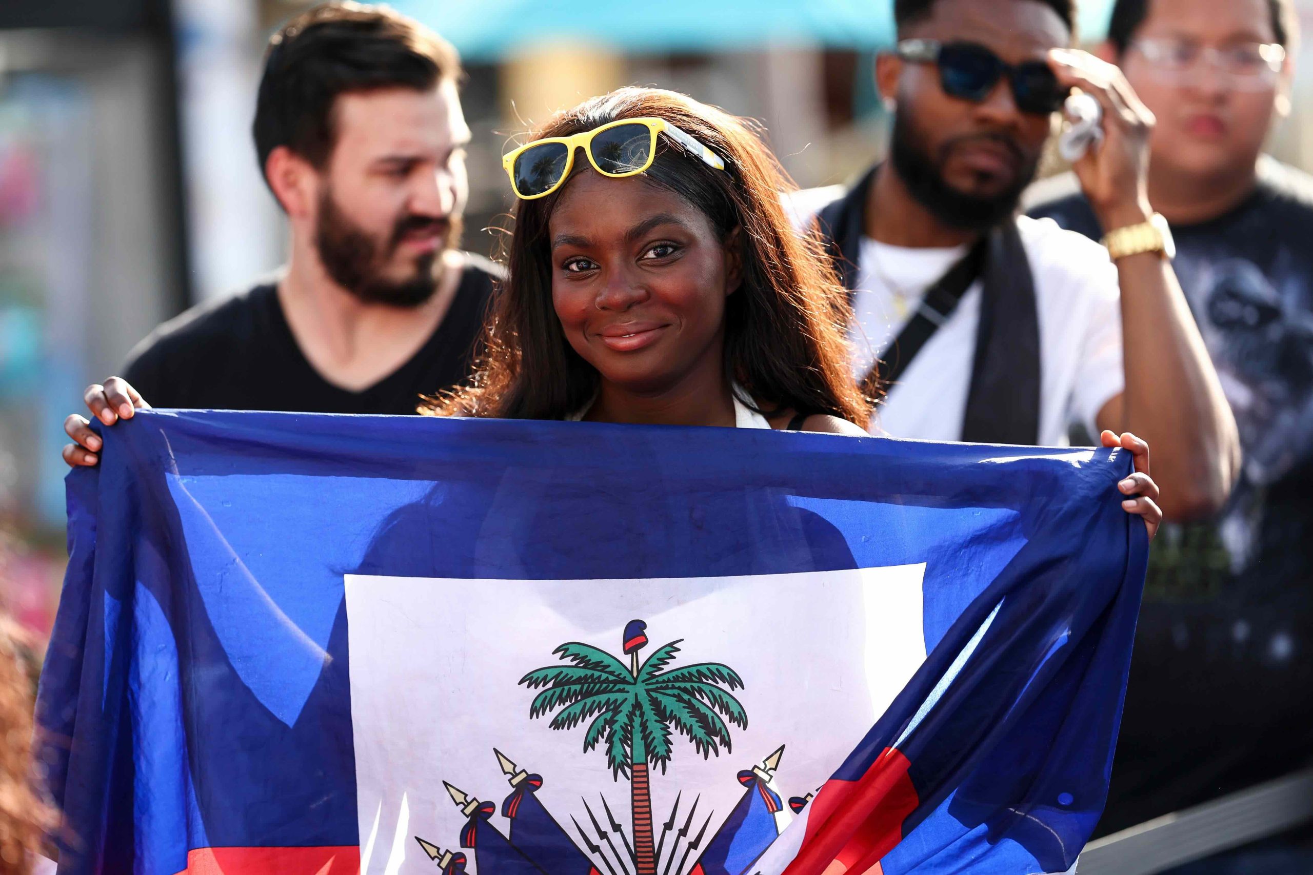 Smiling woman wearing yellow sunglasses holding the Haitian flag at a public event, with people in the background.
