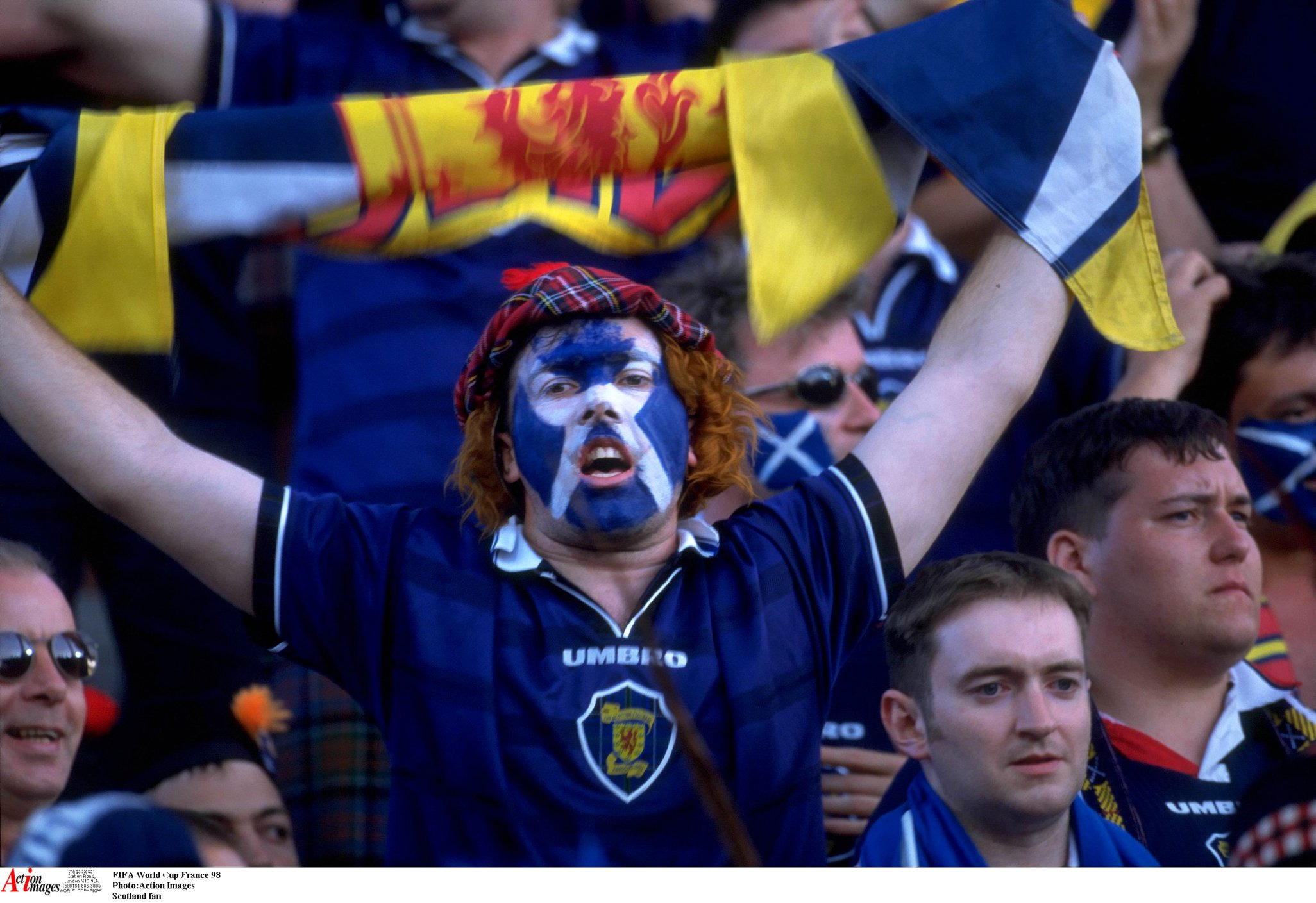 Scotland football fan with face painted in Scottish flag colors, wearing tartan hat, cheering with arms raised and holding a scarf at FIFA World Cup France 1998.