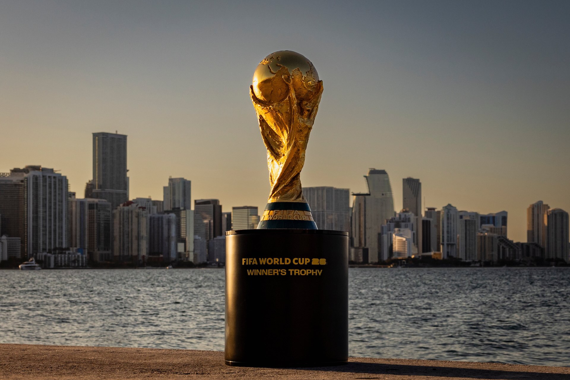 FIFA World Cup Trophy with Miami Skyline at Sunset FIFA World Cup trophy on a pedestal with the Miami skyline in the background at sunset.