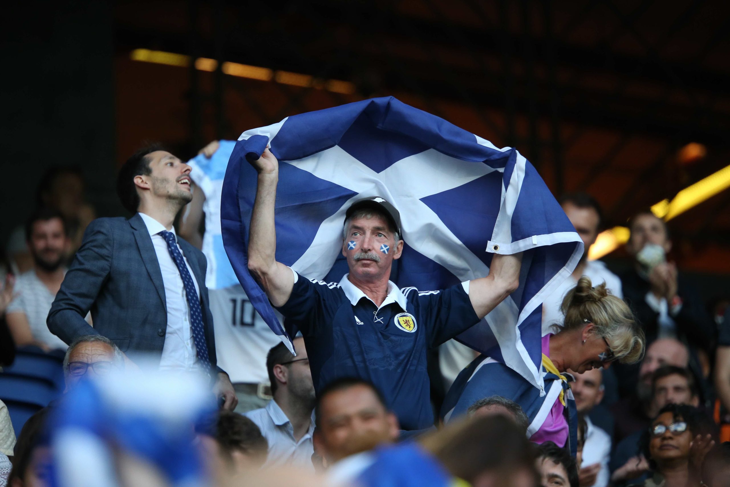 Scotland football fan holding a Saltire flag in a crowded stadium, with face paint and team shirt, cheering during a match.
