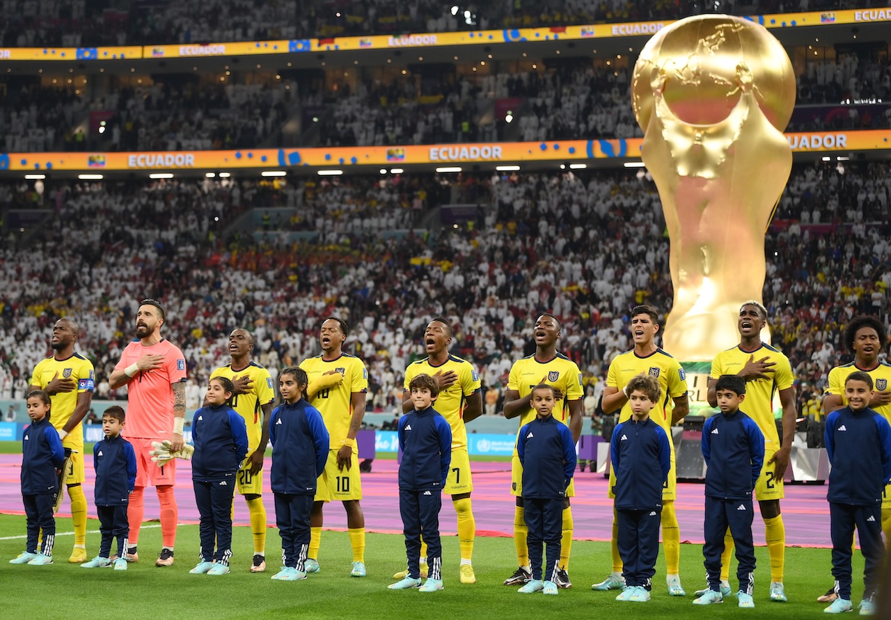Ecuador football players and young mascots sing the national anthem before a World Cup match, with a giant trophy replica behind them.