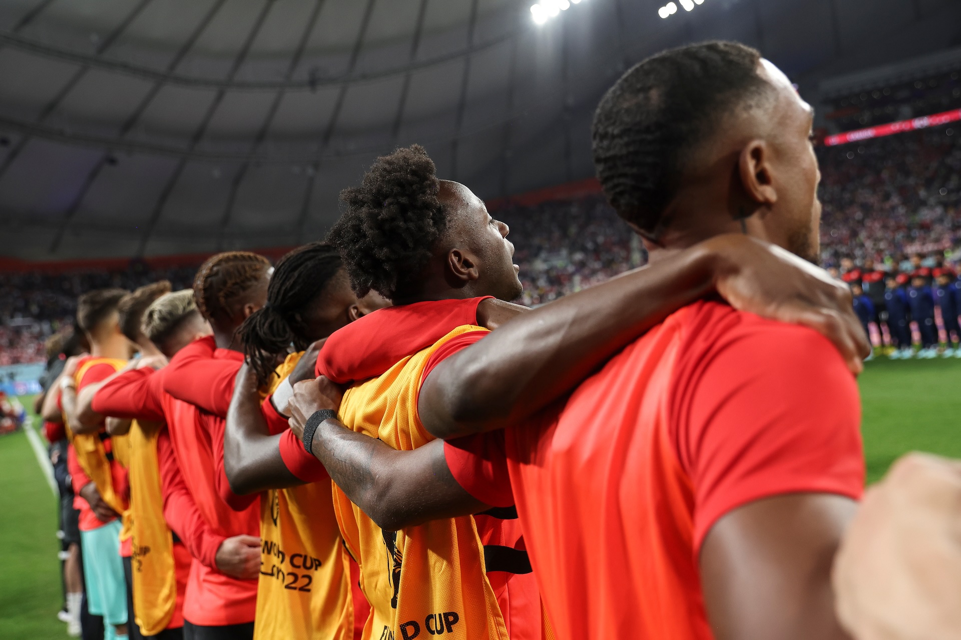 Football players in red and yellow kits standing arm in arm in a huddle before a World Cup match inside a stadium.