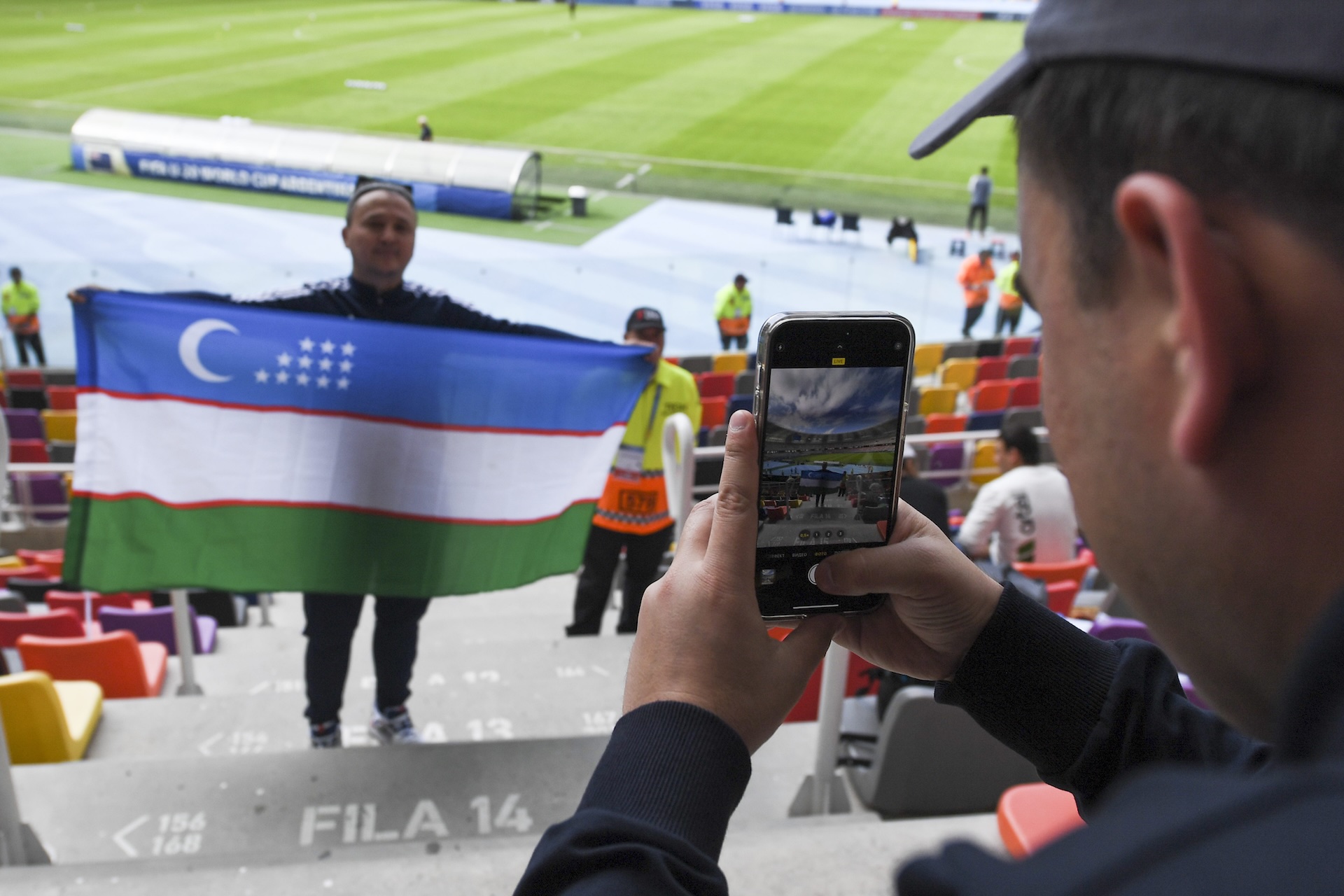 Uzbekistan Football Fan Poses with National Flag in Stadium Uzbekistan football fan holds national flag as another fan takes a photo with a smartphone in a stadium.