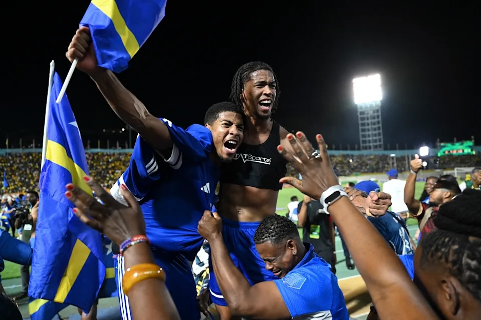 Curaçao football players and fans celebrate together on the field, waving flags and cheering after a victory.