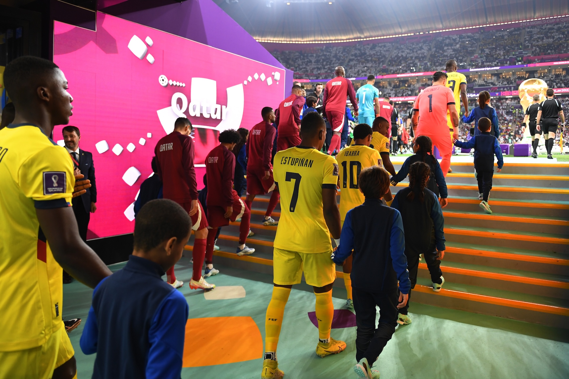 Ecuador and Qatar football players, with young mascots, walk up stadium steps to enter the field for a World Cup match.