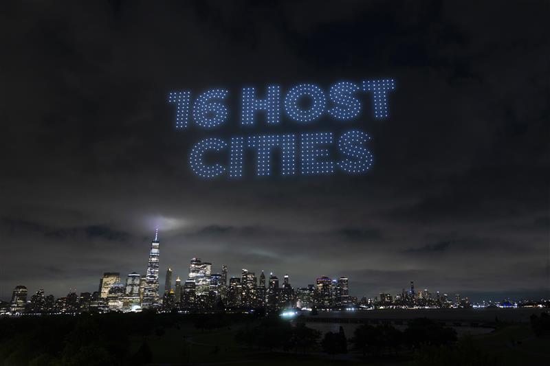 New York City skyline at night with "16 Host Cities" written in the sky by a drone light display.