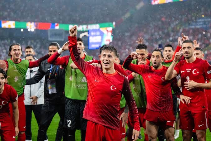 Turkey national football team players in red kits celebrate on the field after a match, smiling and raising their arms.